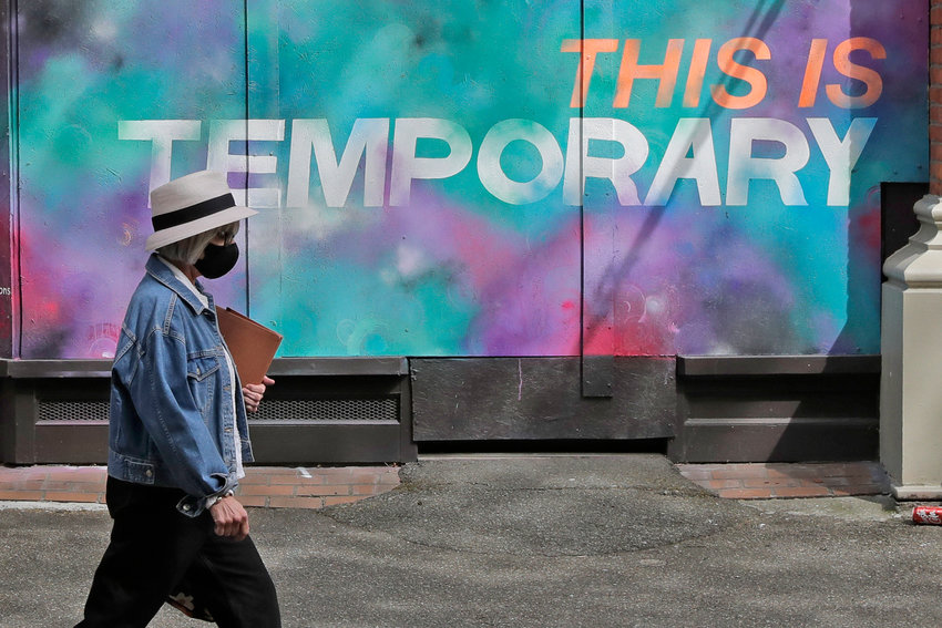 A person wearing a mask walks past a mural on a boarded-up window that reads 'This is temporary.'