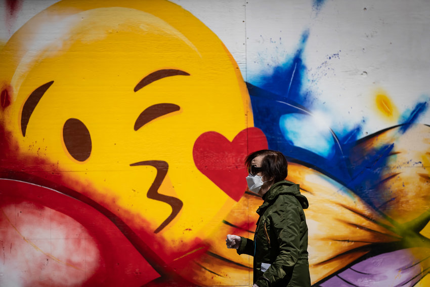 A man in a mask walks past a mural with a giant emoji.