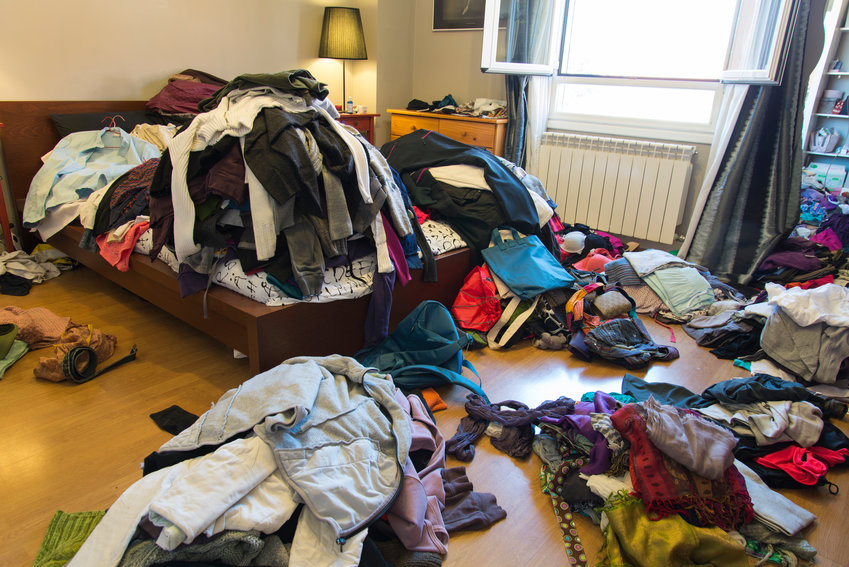 A room with clothes on the floor and piled high on the bed.